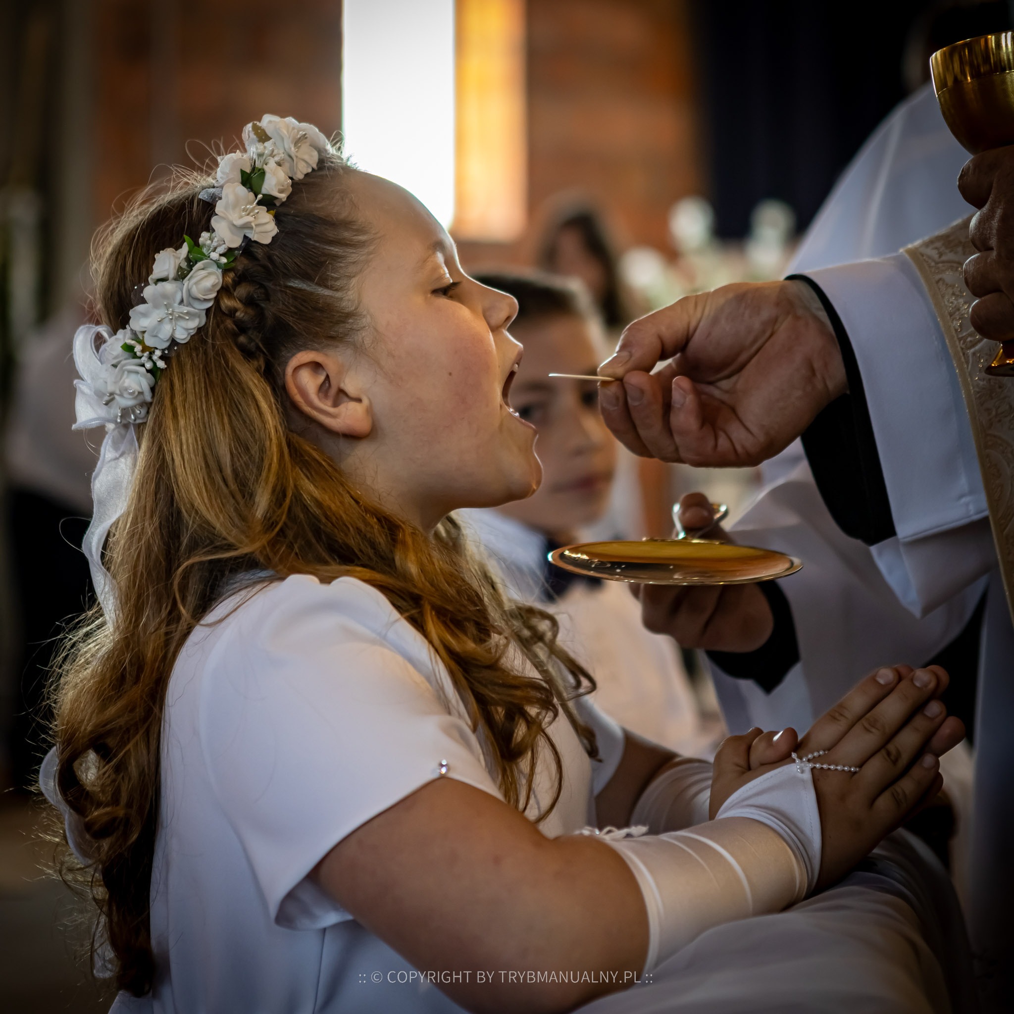 Dziewczynka w białej sukience i wianku przyjmuje komunię świętą, z rękami złożonymi do modlitwy, w tle widoczny ksiądz z kielichem i inni uczestnicy uroczystości.