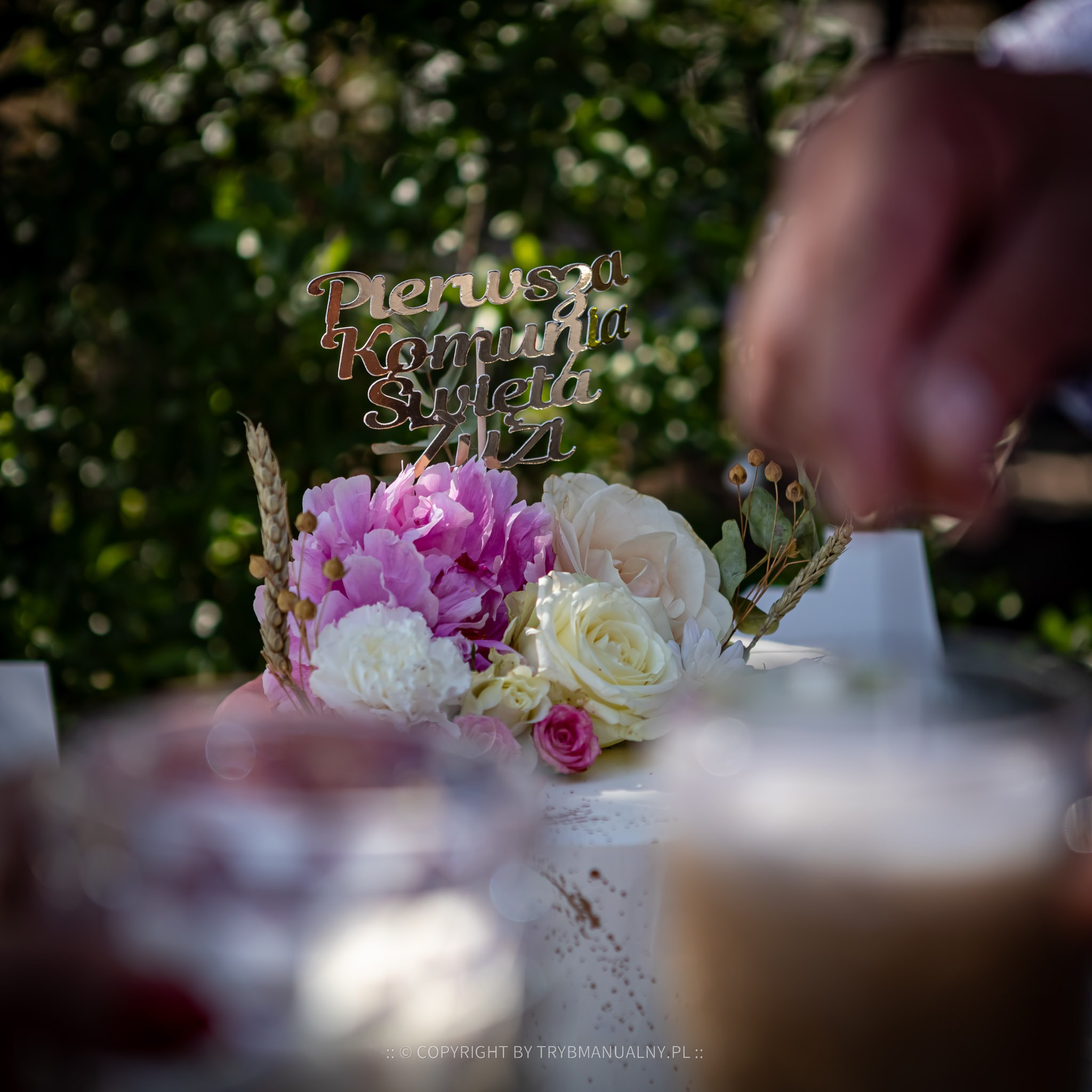 Tort z okazji Pierwszej Komunii Świętej udekorowany kwiatami i złotym napisem 'Pierwsza Komunia Święta 2021', sfotografowany w Warszawie z perspektywy osoby trzymającej kieliszek.