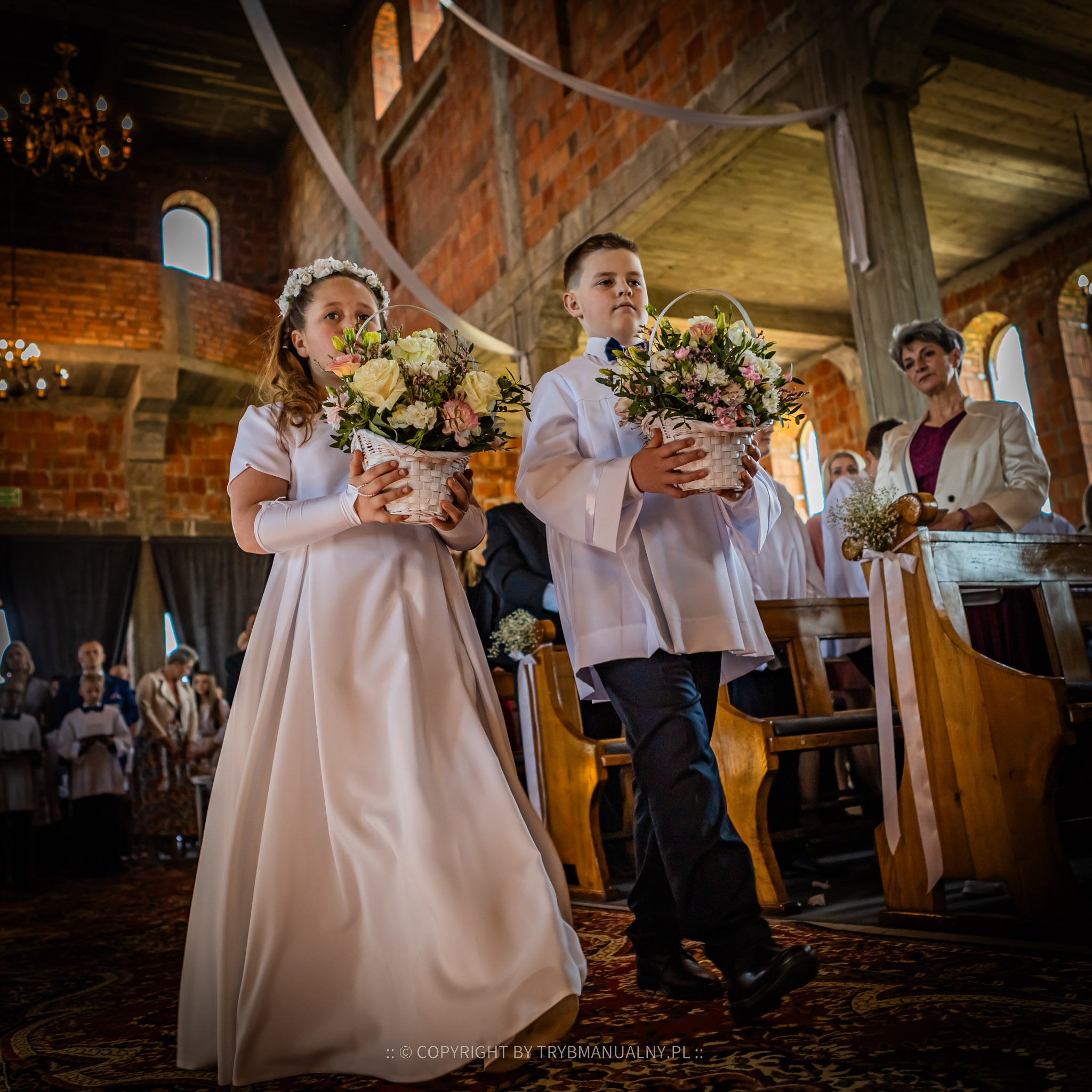 Dwoje dzieci w strojach komunijnych niesie koszyki z kwiatami w kościele z ceglanymi ścianami i drewnianymi ławkami, w tle widoczni goście.