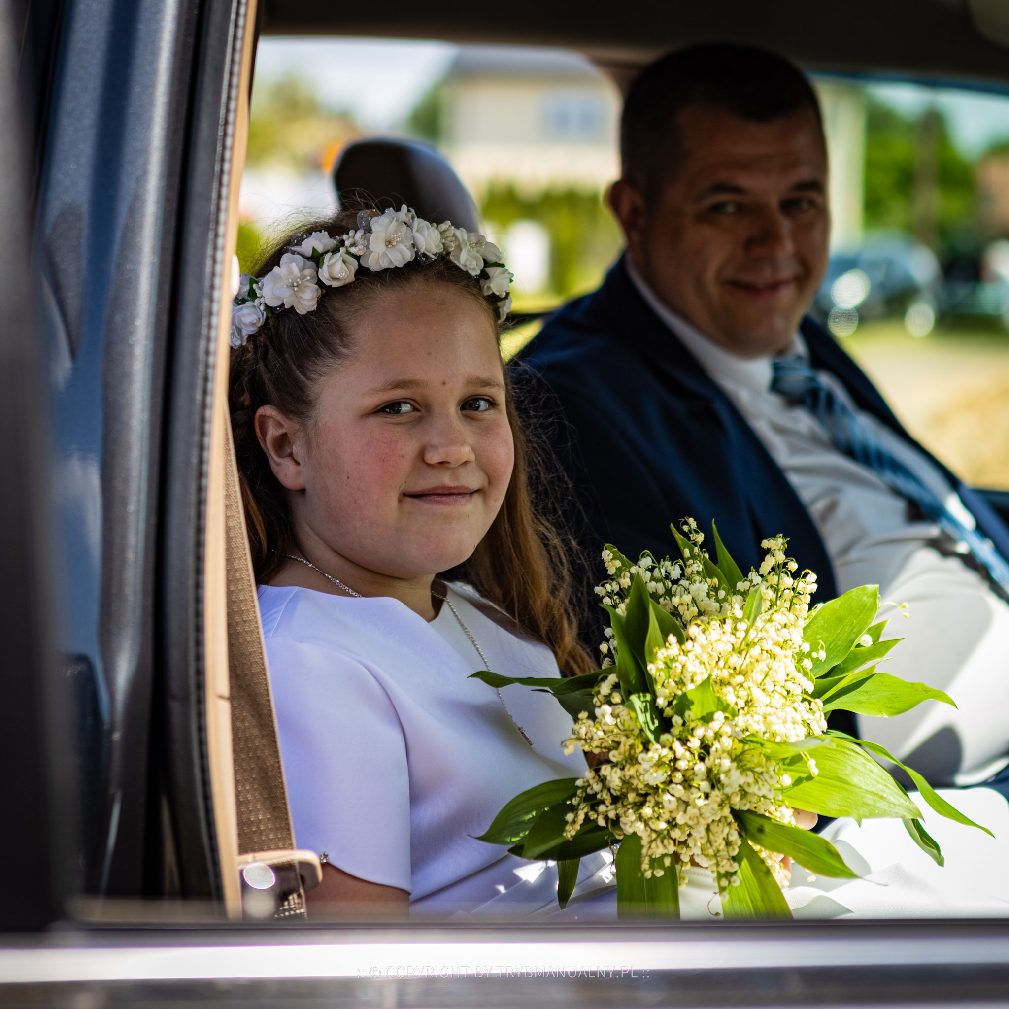 Dziewczynka w wianku z białych kwiatów i białej sukience, siedząca w samochodzie z bukietem konwalii, obok mężczyzna w garniturze.