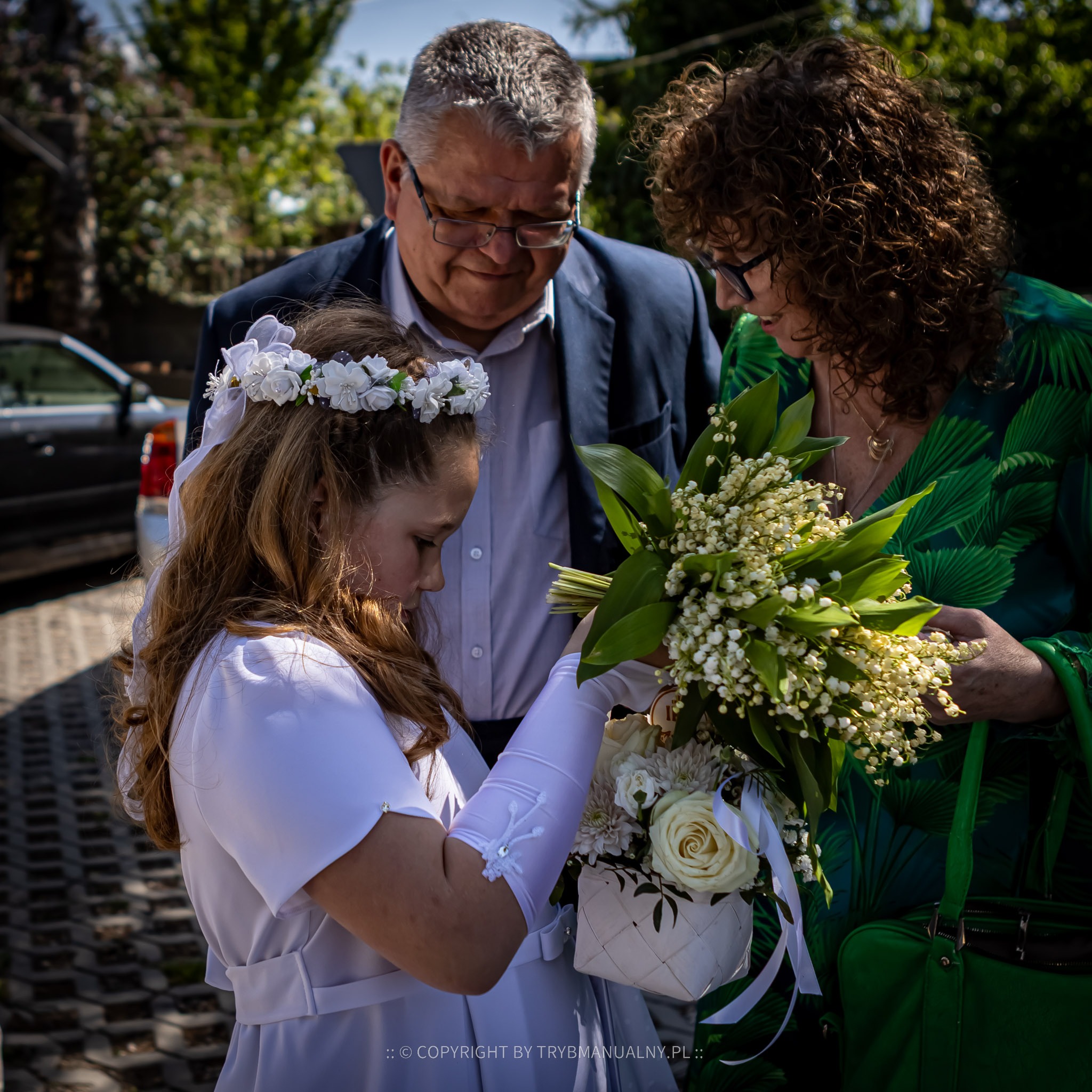 Dziewczynka w białej sukience komunijnej z wiankiem na głowie trzyma bukiet konwalii i białe róże, obok rodzice obserwujący z uśmiechem.