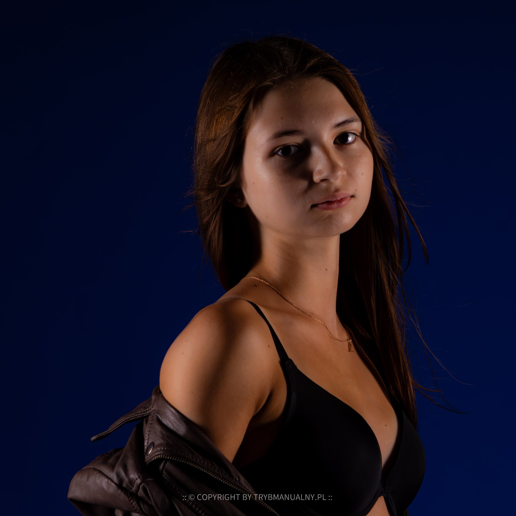 Portret młodej kobiety w czarnej bieliźnie i skórzanej kurtce na tle ciemnoniebieskiego tła, włosy rozwiane
