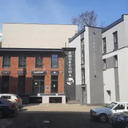 Widok zewnętrzny budynku usługowego z ceglaną fasadą i napisem 'Dworcowa 45' oraz nowoczesnego budynku apartamentowego w szarościach, z zaparkowanymi samochodami na pierwszym planie.