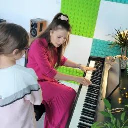 Kobieta w różowej sukience demonstruje grę na keyboardzie dziewczynce w pokoju z panelami akustycznymi i rośliną doniczkową.