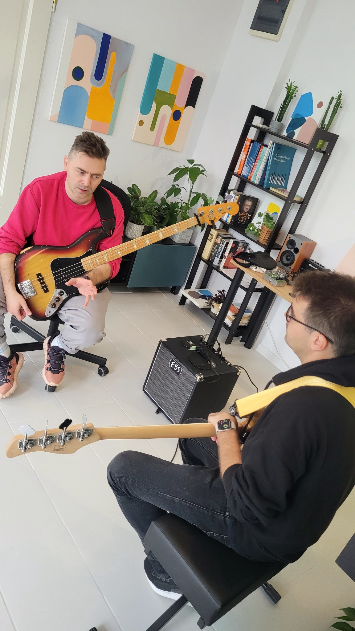 Dwóch muzyków siedzi na krzesłach i gra na gitarach basowych w jasnym pokoju z roślinami i półką na książki.