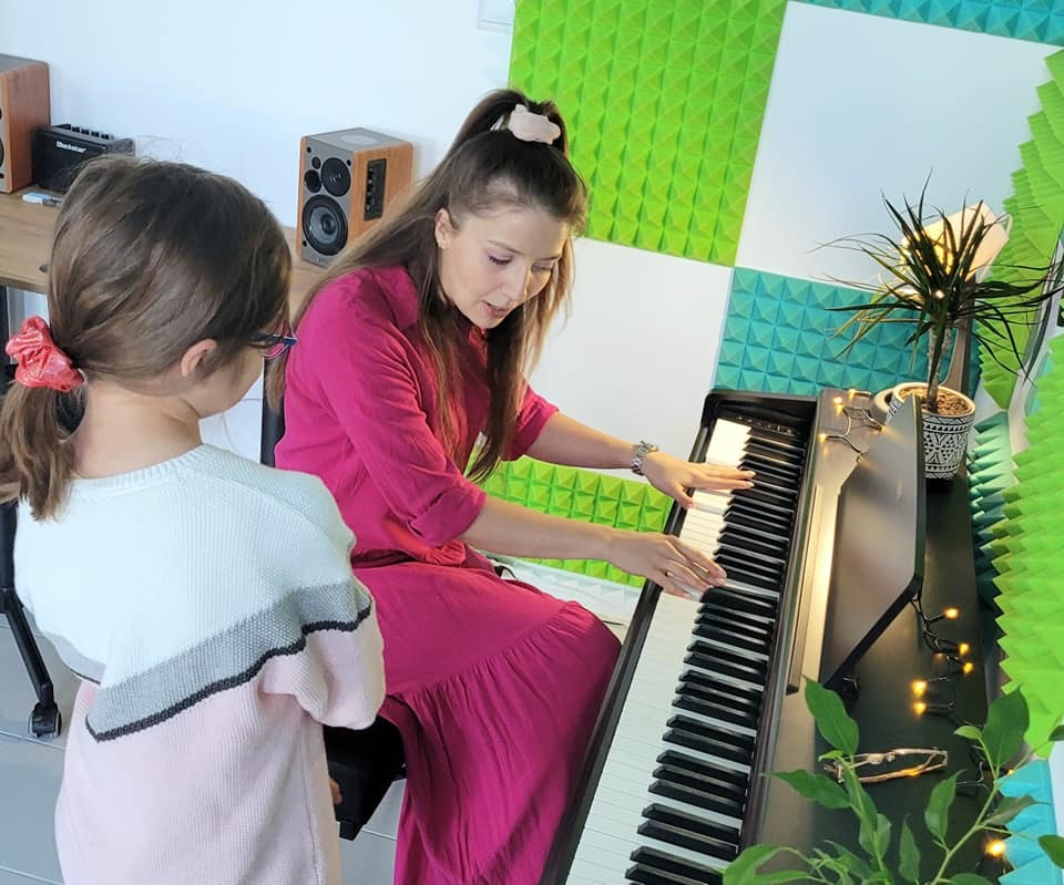 Kobieta w różowej sukience demonstruje grę na keyboardzie dziewczynce w pokoju z panelami akustycznymi i rośliną doniczkową.