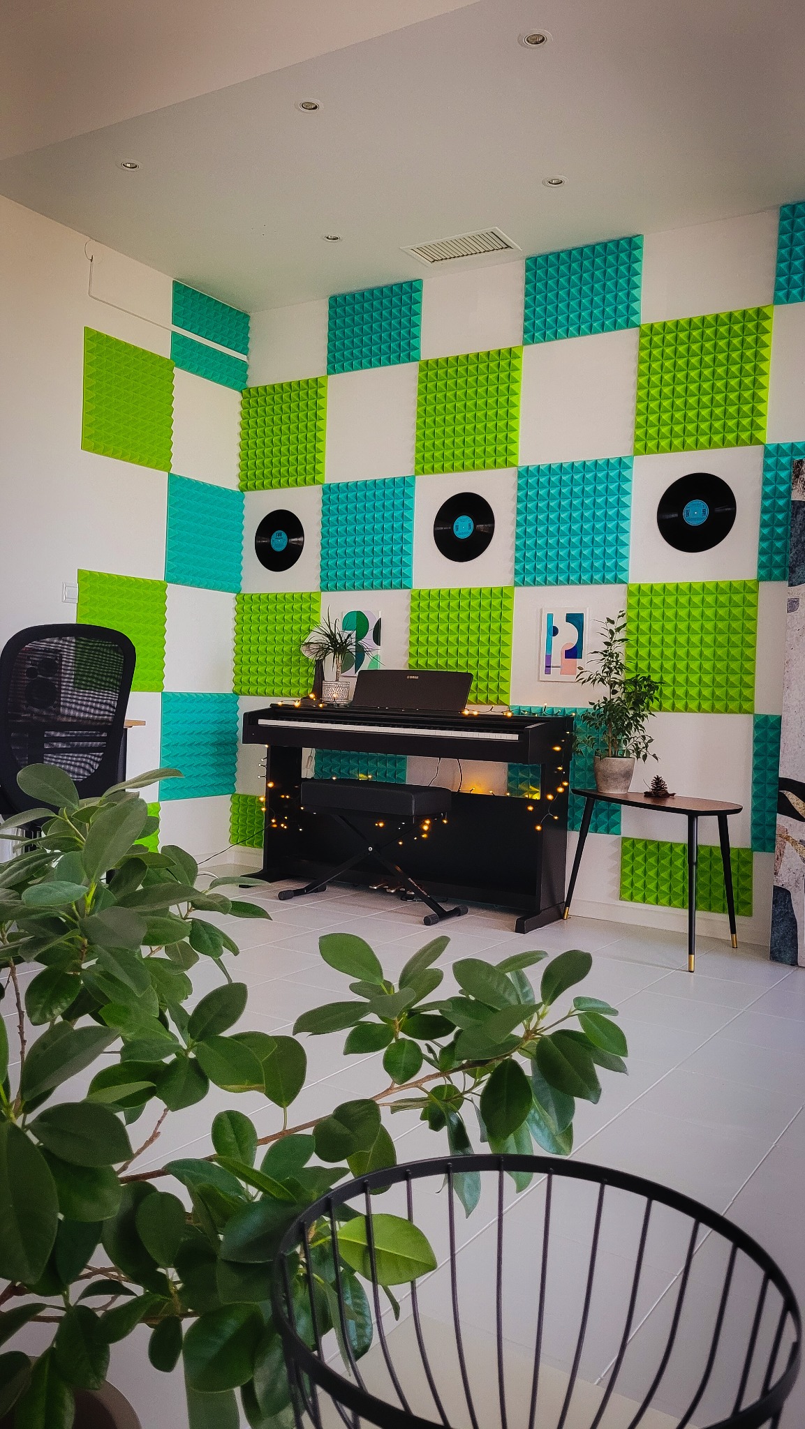 Pokój z pianinem cyfrowym Yamaha, dekoracją ścian z paneli akustycznych w szachownicę (zielone, turkusowe i białe) oraz płytami winylowymi, oświetlony lampkami.