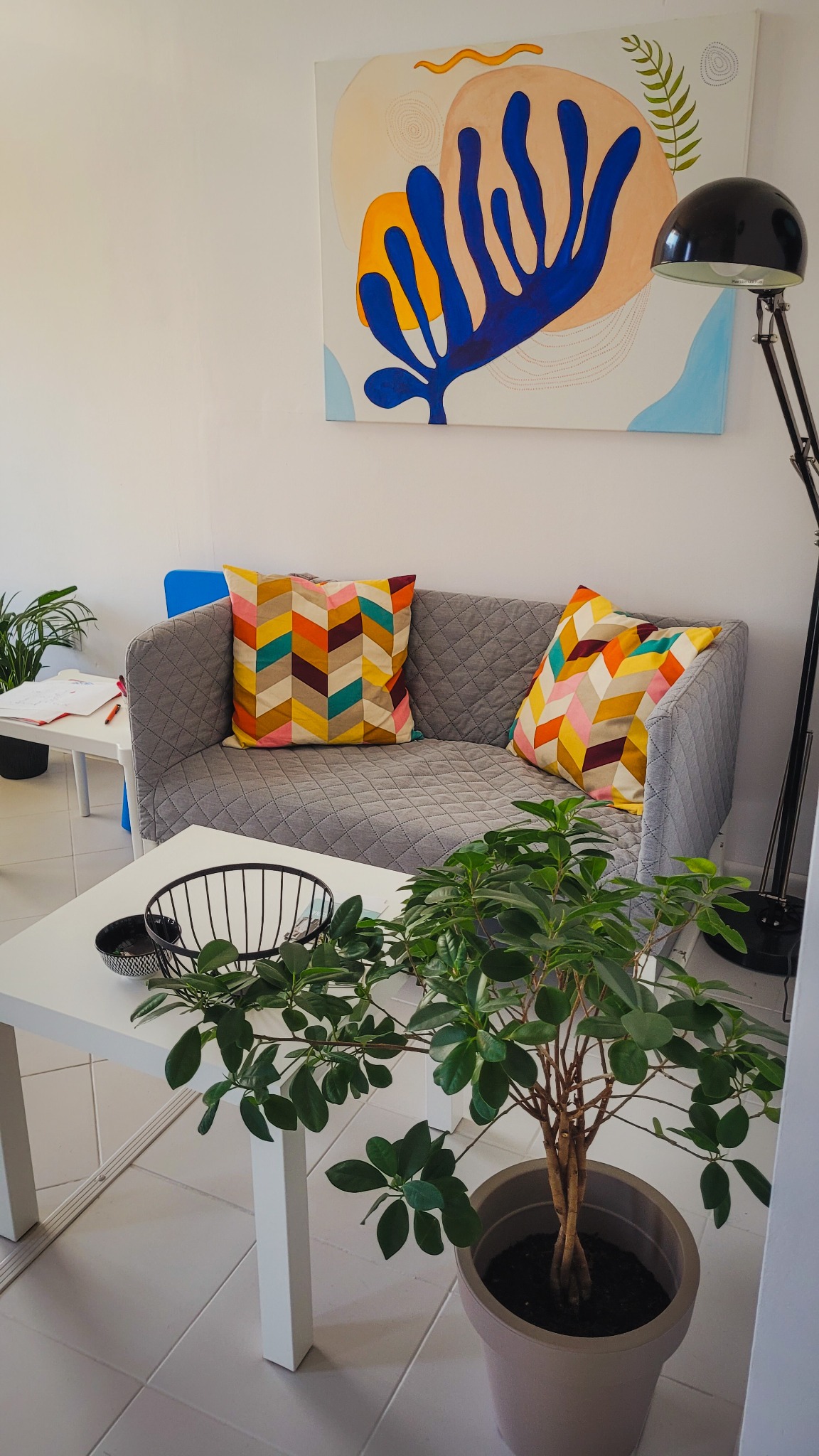 Szary tapicerowany narożnik z geometrycznymi poduszkami, abstrakcyjny obraz na ścianie i roślina doniczkowa na białym stoliku kawowym, oświetlone czarną lampą podłogową.