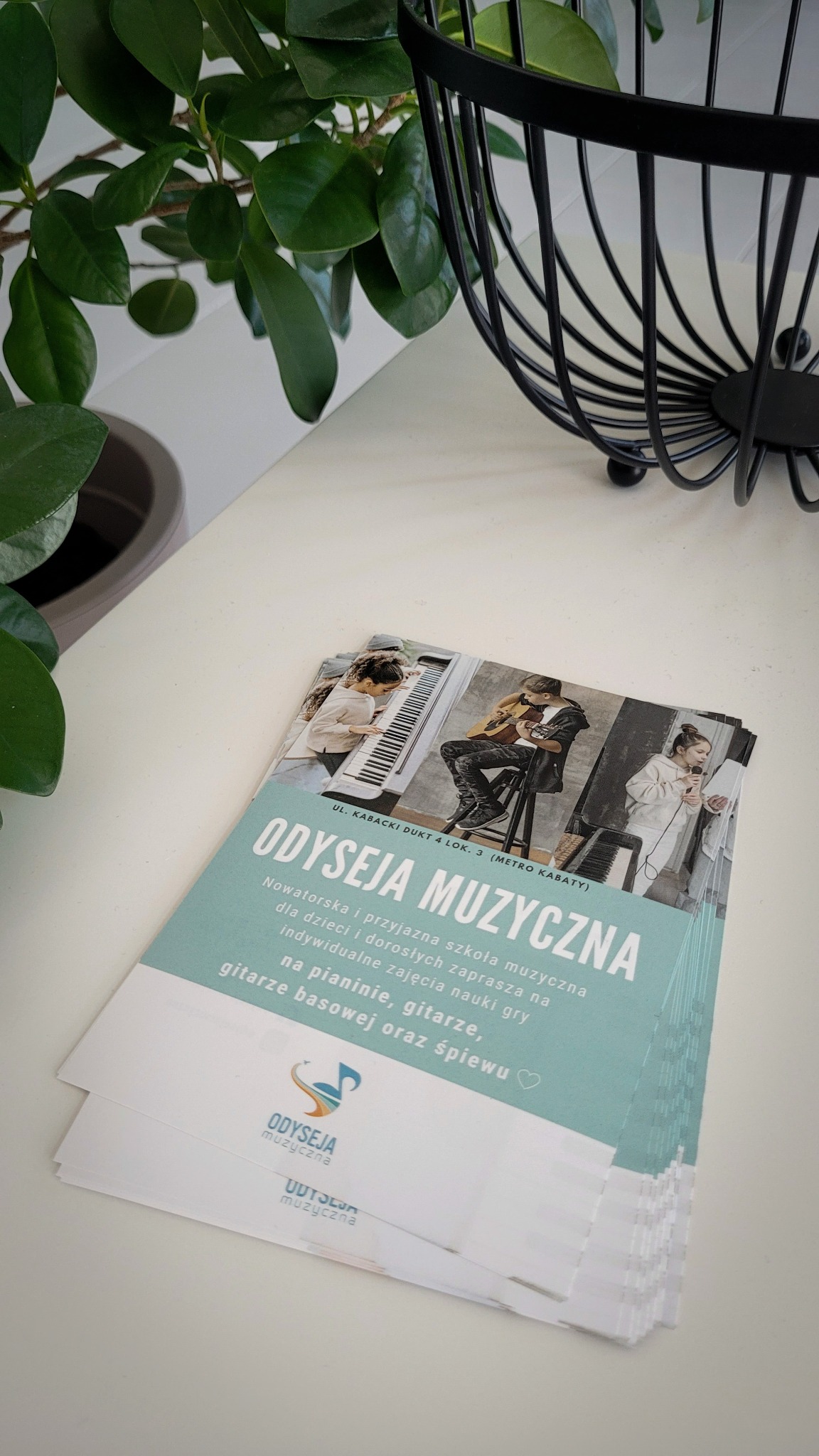 Stos ulotek reklamujących szkołę muzyczną 'Odyseja Muzyczna' z ofertą nauki gry na pianinie, gitarze, gitarze basowej i śpiewu, leżący na białym stole obok czarnego koszyka i rośliny doniczkowej.