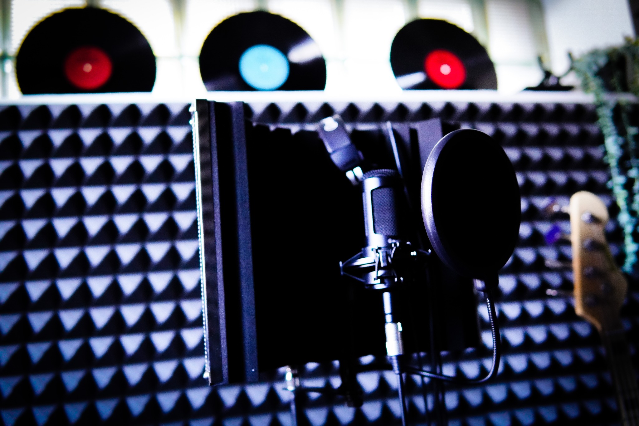 Mikrofon studyjny z filtrem pop i ekranem akustycznym na tle pianki akustycznej oraz winyli i części gitary basowej.