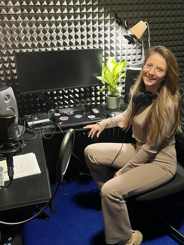Uśmiechnięta kobieta w okularach i słuchawkach, siedząca w studiu nagraniowym w Warszawie, z mikrofonem i monitorem na biurku, na tle akustycznych paneli ściennych.
