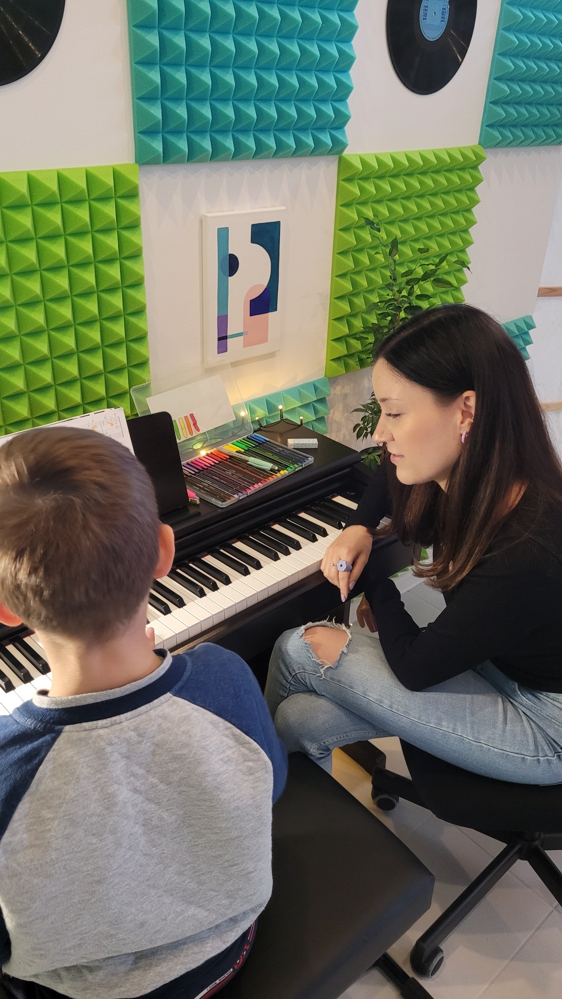 Chłopiec uczy się grać na pianinie pod okiem instruktorki w pokoju z akustycznymi panelami; na klawiaturze leżą kolorowe pisaki.