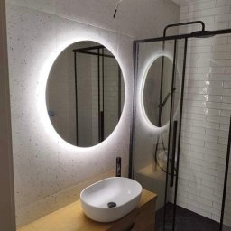 KG-Remonty wykończenia wnętrz Krzysztof Górowski - Nowoczesna łazienka z okrągłym lustrem LED, umywalką nablatową na drewnianej szafce i kabiną prysznicową z czarnymi profilami. Minimalistyczny design i jasne płytki.