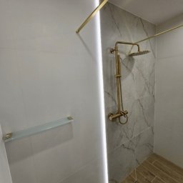 KG-Remonty wykończenia wnętrz Krzysztof Górowski - Wykończona kabina prysznicowa z marmurowymi płytkami, złotą armaturą i szklaną półką. Nowoczesny design z oświetleniem LED wzdłuż krawędzi. Drewnopodobna podłoga.