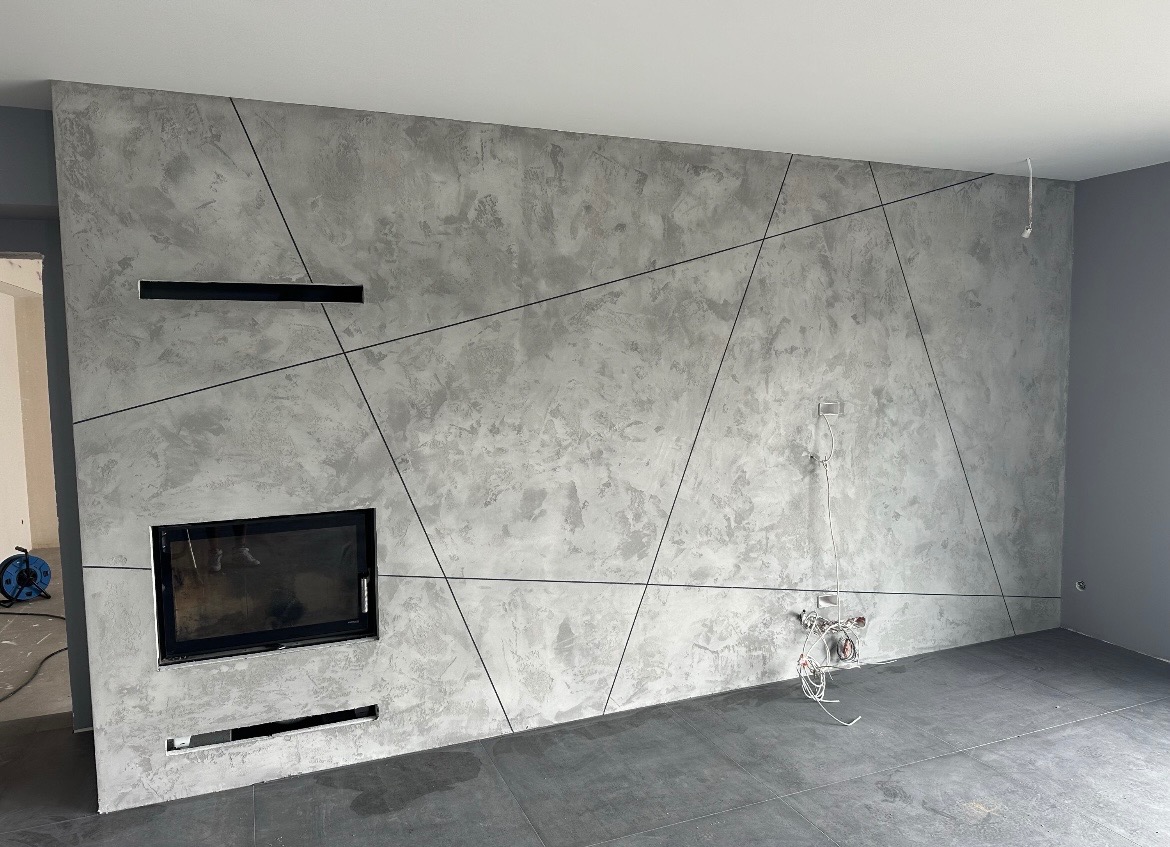 Ściana z szarą, dekoracyjną fakturą i geometrycznymi liniami, wbudowany kominek, szara podłoga. Widoczne przewody elektryczne. Nowoczesne wykończenie wnętrza.