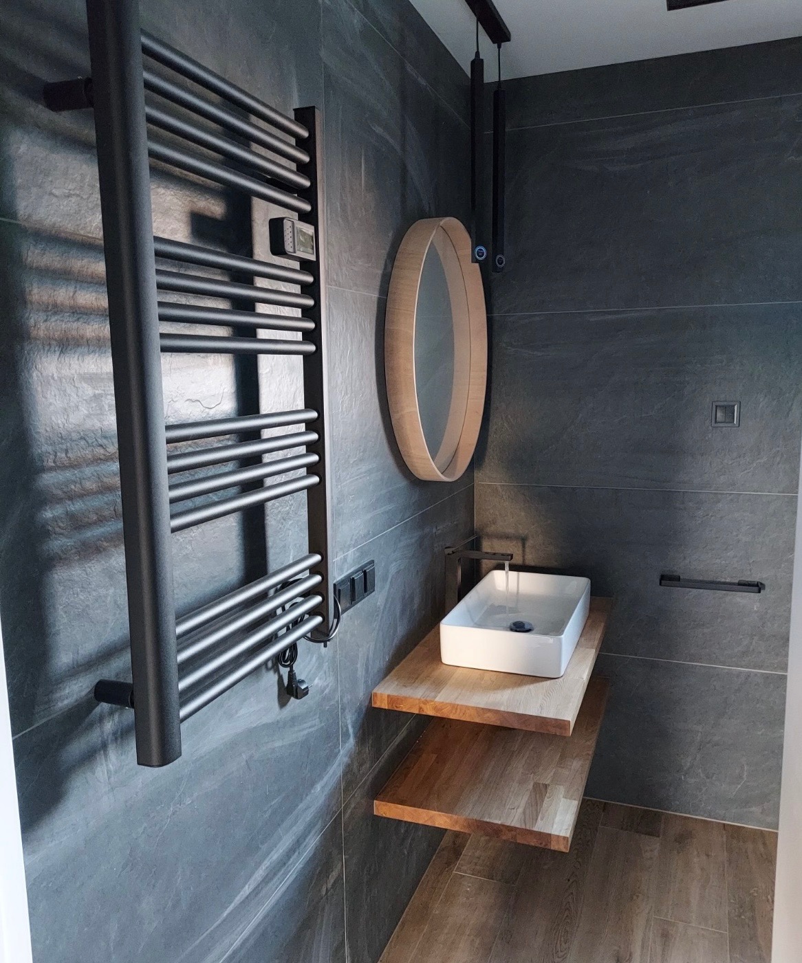 Nowoczesna łazienka z szarymi płytkami, drewnianymi półkami pod umywalką i owalnym lustrem. Czarny grzejnik drabinkowy i minimalistyczna armatura.