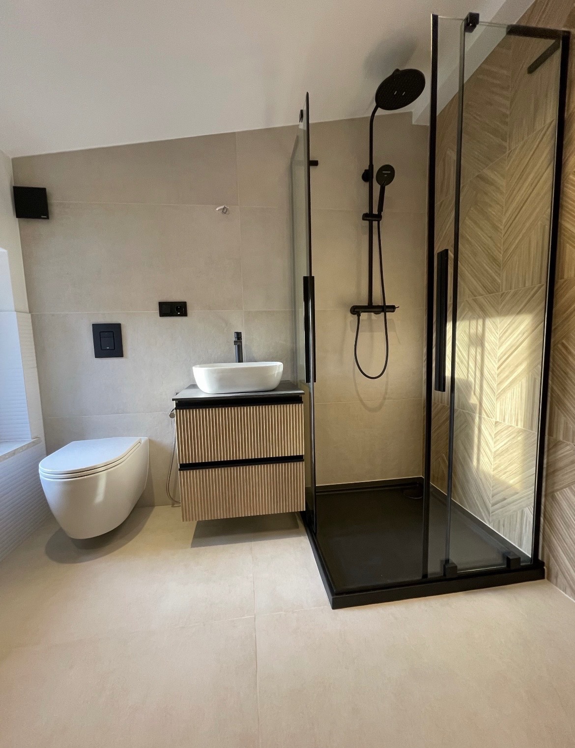 Nowoczesna łazienka z beżowymi płytkami, kabiną prysznicową typu walk-in z czarnymi elementami i umywalką nablatową na szafce z lamelami. Minimalistyczny design.