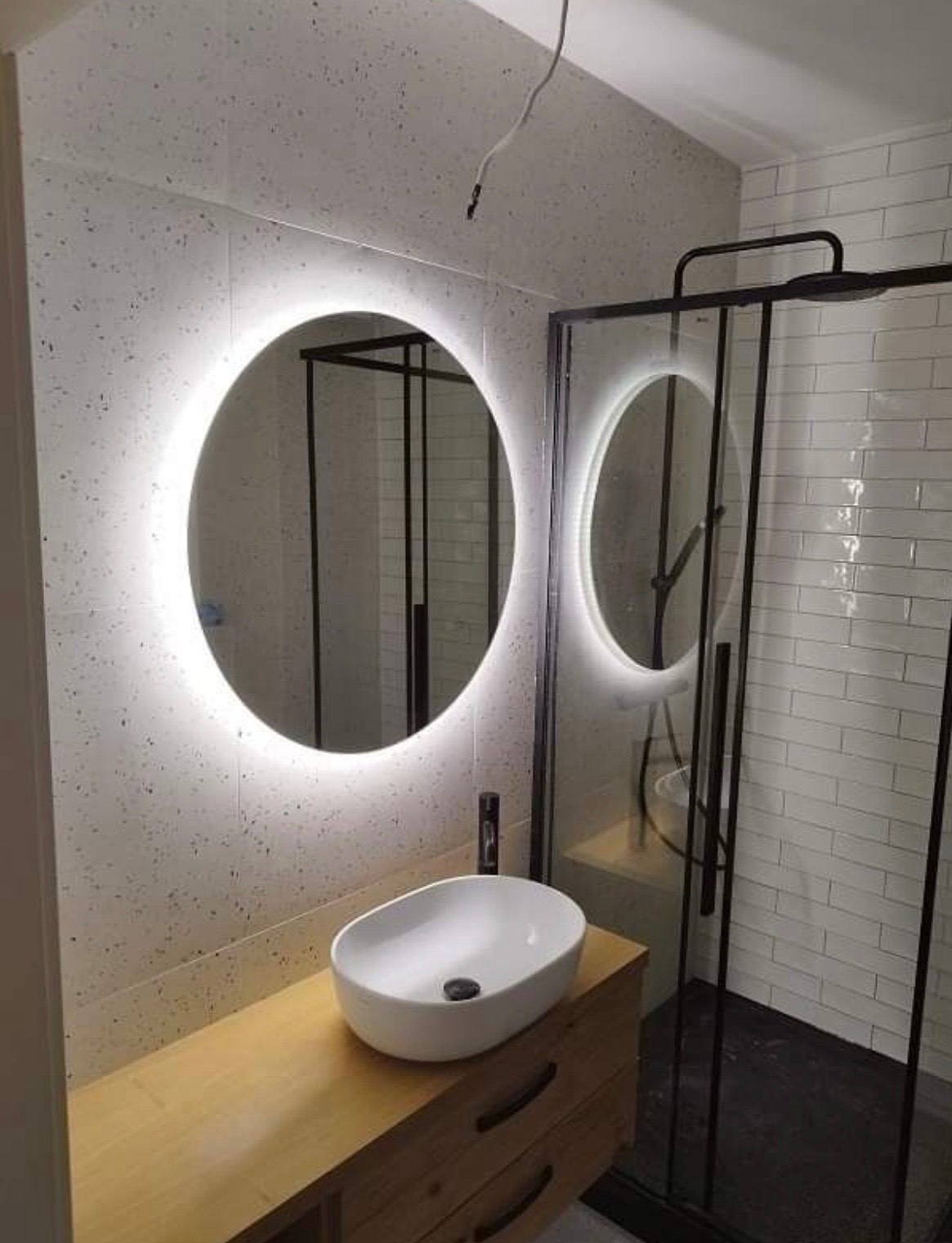 Nowoczesna łazienka z okrągłym lustrem LED, umywalką nablatową na drewnianej szafce i kabiną prysznicową z czarnymi profilami. Minimalistyczny design i jasne płytki.