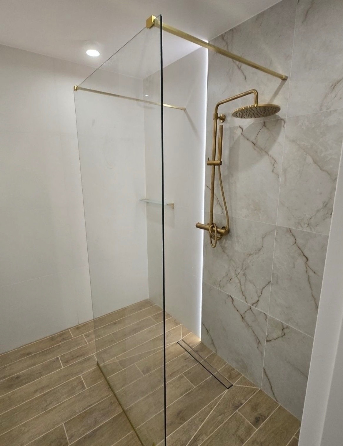 Kabina prysznicowa walk-in z złotą armaturą i odpływem liniowym, wykończona płytkami imitującymi marmur i drewno. Realizacja w Nowym Sączu.
