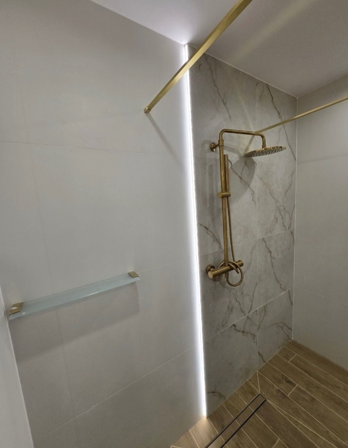 Wykończona kabina prysznicowa z marmurowymi płytkami, złotą armaturą i szklaną półką. Nowoczesny design z oświetleniem LED wzdłuż krawędzi. Drewnopodobna podłoga.