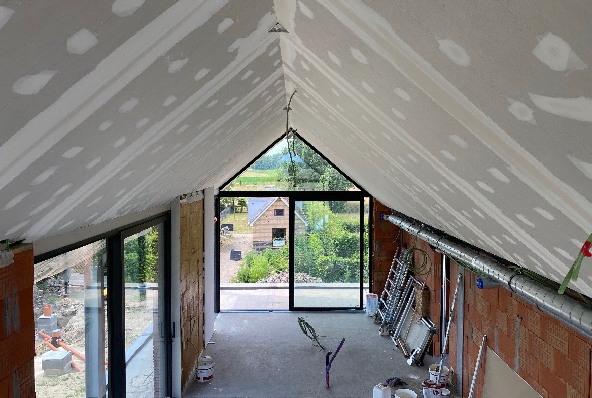 Poddasze w trakcie prac wykończeniowych: ściany z cegły, okna z widokiem na zieleń, sufit z nałożoną gładzią, widoczne narzędzia i materiały budowlane.