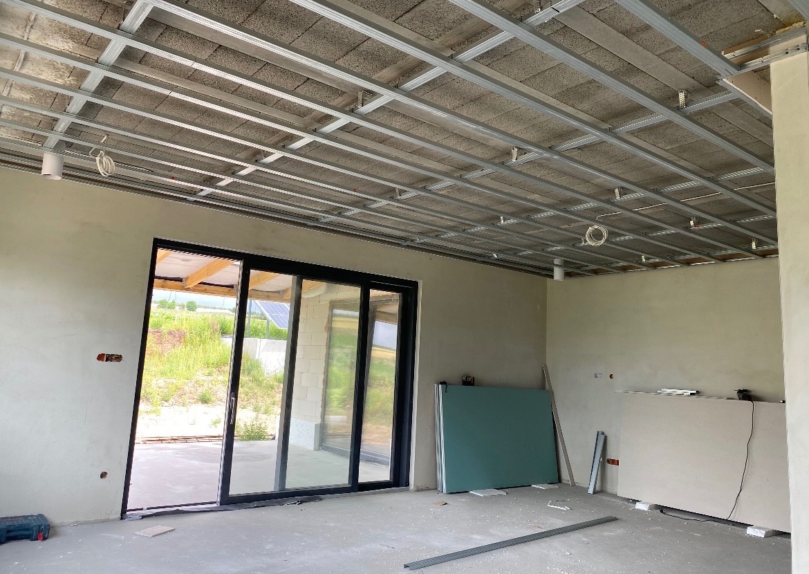 Wnętrze w trakcie prac wykończeniowych: metalowa konstrukcja sufitu podwieszanego, ściany po gładziowaniu, duże okno przesuwne z widokiem na zewnątrz, płyty gipsowe.
