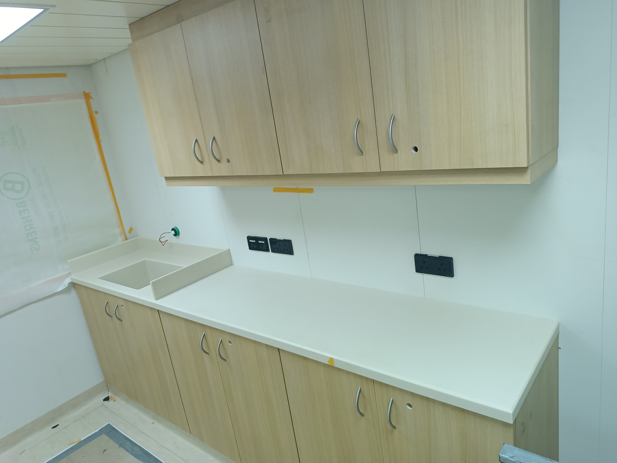 Jasne, drewniane szafki kuchenne z białym blatem i zlewozmywakiem w Gorzowie Wielkopolskim, widoczne gniazdka elektryczne na ścianie.
