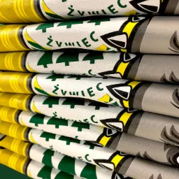 Stos zwiniętych, zadrukowanych materiałów z motywami graficznymi, w tym stylizowane drzewa i wizerunek wilka, w kolorach zielonym, żółtym, białym i szarym.