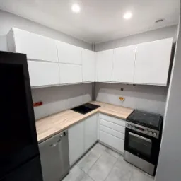 Nowoczesna, narożna kuchnia z białymi, gładkimi frontami szafek, jasnym blatem imitującym drewno i czarnym zlewem, widziana z perspektywy lekko z góry, z widoczną czarną lodówką po lewej.