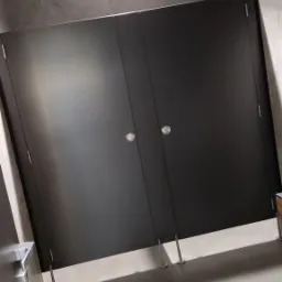 Ciemne, dwuskrzydłowe drzwi do kabiny sanitarnej z metalowymi uchwytami i nóżkami, widoczne na tle szarej ściany i podłogi z płytek.