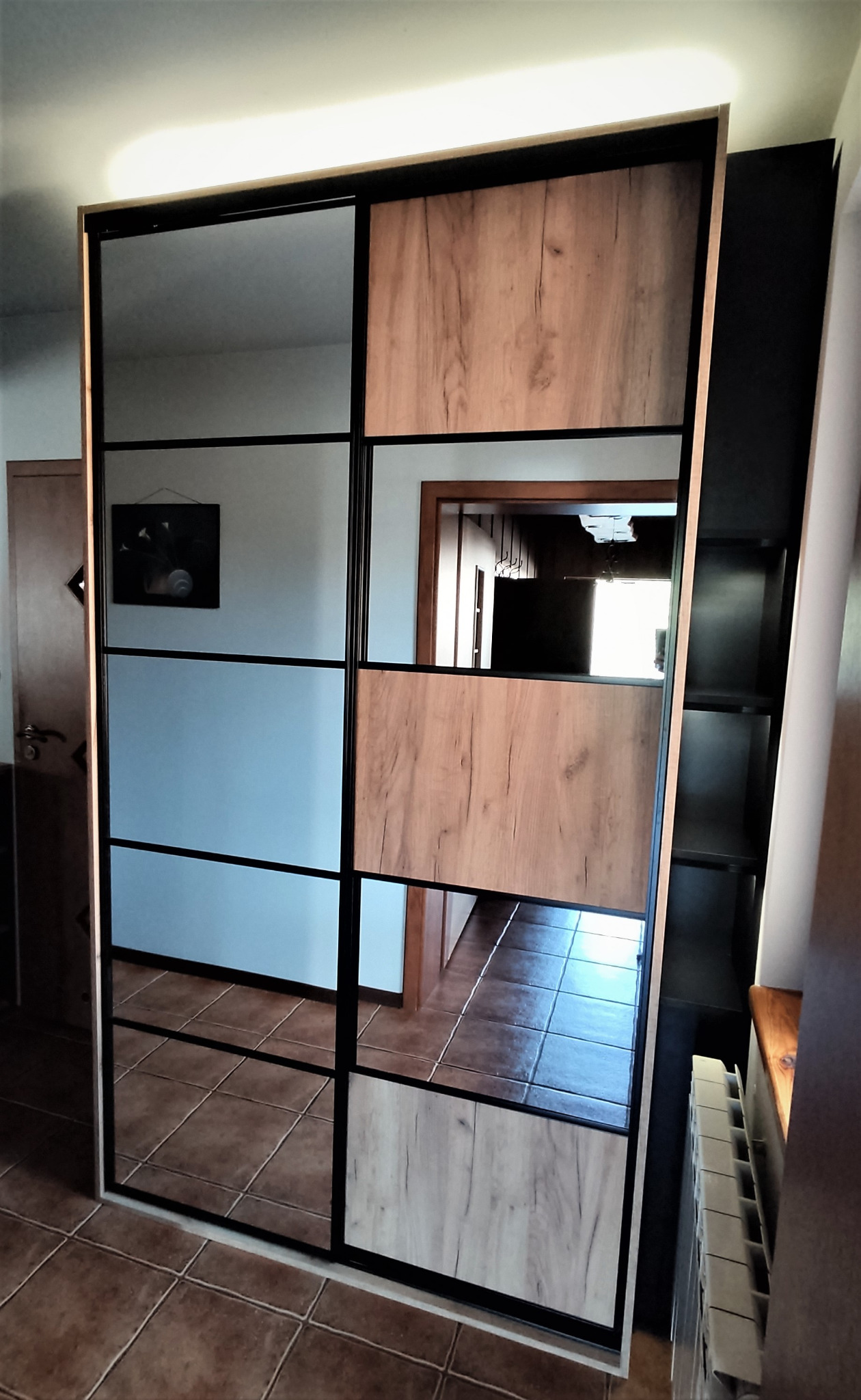 Szafa wnękowa z przesuwnymi drzwiami, czarnymi ramami i panelami z imitacją drewna oraz lustrzanymi wstawkami, odbijająca fragment wnętrza z brązowymi płytkami podłogowymi.