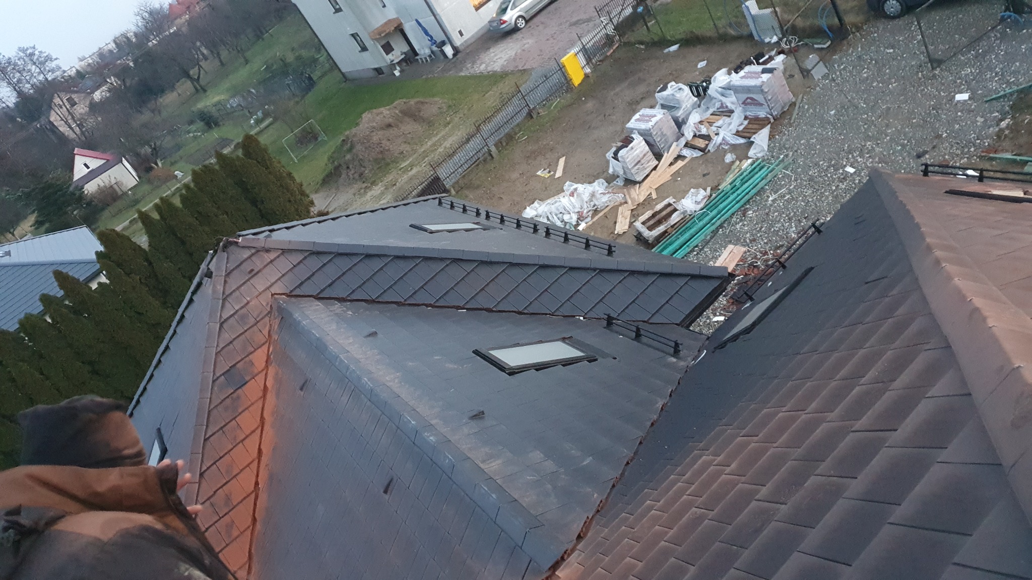 Nowy dach z dachówką ceramiczną w kolorze grafitowym, z oknami połaciowymi, widok z góry, materiały budowlane składowane na posesji.