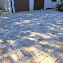 Ułożona kostka brukowa w odcieniach szarości i beżu, tworząca wzór na podjeździe przed domem z dwoma garażami, z widocznymi cieniami drzew.