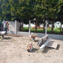 CZU-BART - Montaż ogrodzenia panelowego z betonową podmurówką. Widoczne słupki, panele, taczki i piła do betonu. W tle rząd drzew.