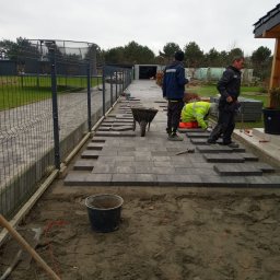 CZU-BART - Trzech pracowników układa szarą kostkę brukową na posesji, widoczny fragment ogrodzenia, taczka i narzędzia. Częściowo ułożony chodnik prowadzi w głąb działki.