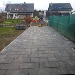 CZU-BART - Układanie szarej kostki brukowej na posesji. Widoczni dwaj pracownicy, jeden z zagęszczarką. W tle domy jednorodzinne i zielona siatka ogrodzeniowa.