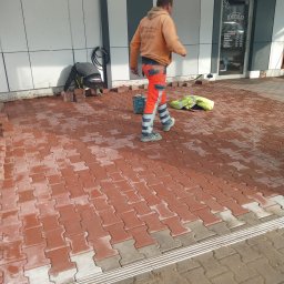 CZU-BART - Układanie czerwonej kostki brukowej typu I na zewnątrz budynku. Dwóch pracowników w odblaskowych ubraniach podczas pracy. Częściowo ułożona nawierzchnia.