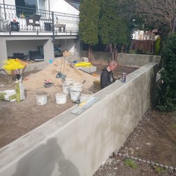 CZU-BART - Pracownik wygładza świeżo wylaną, betonową ścianę oporową w ogrodzie. Widoczne narzędzia, worki z zaprawą, piasek i dom w tle. Praca w toku, etap wykończeniowy.