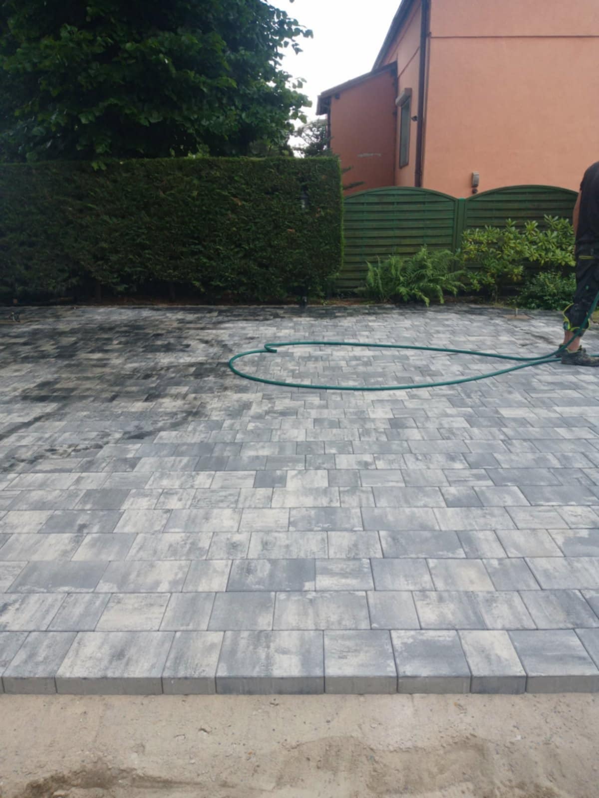Ułożona kostka brukowa w odcieniach szarości, częściowo mokra. W tle zielony żywopłot, drewniany płot i fragment budynku. Wąż ogrodowy na kostce.