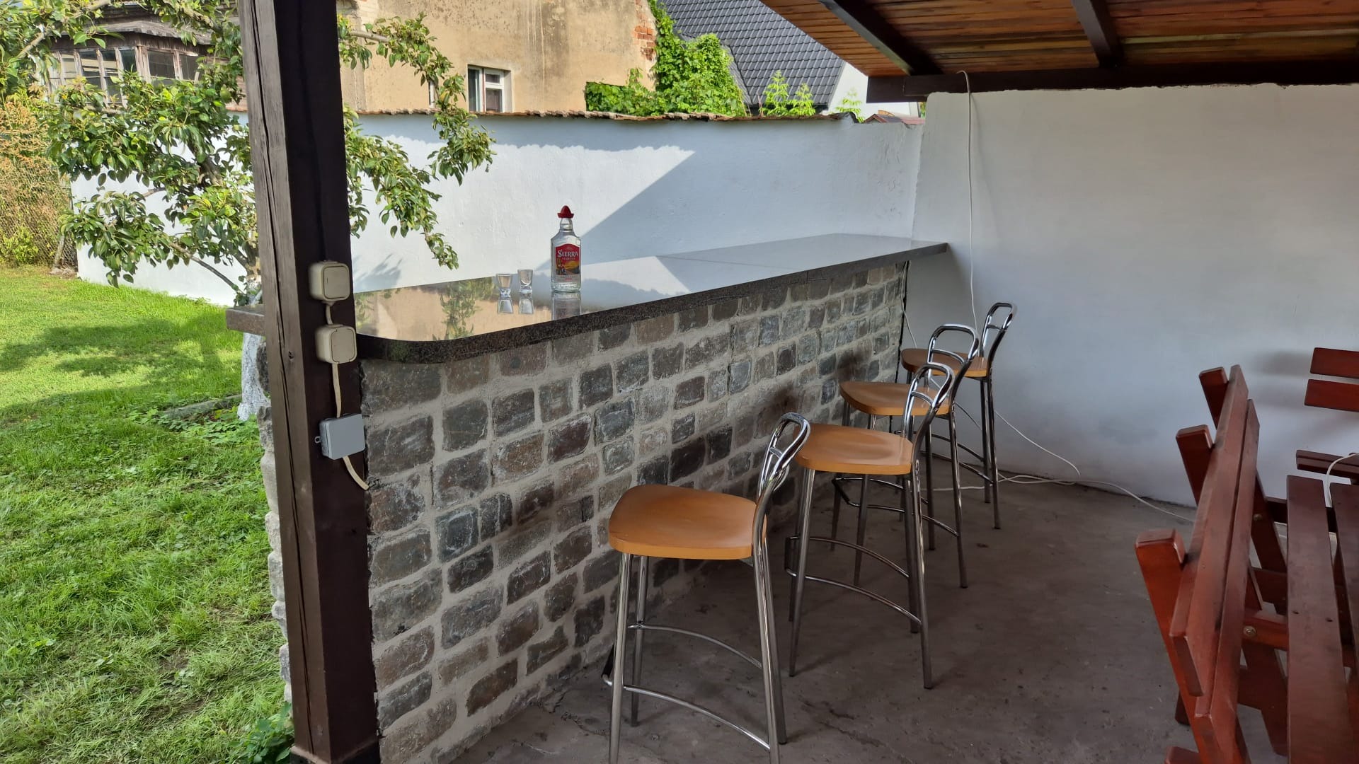 Zadaszony bar ogrodowy z kamienną ścianą, blatem z ciemnego granitu, trzema krzesłami barowymi i butelką tequili. Widok na trawnik i budynek w tle.