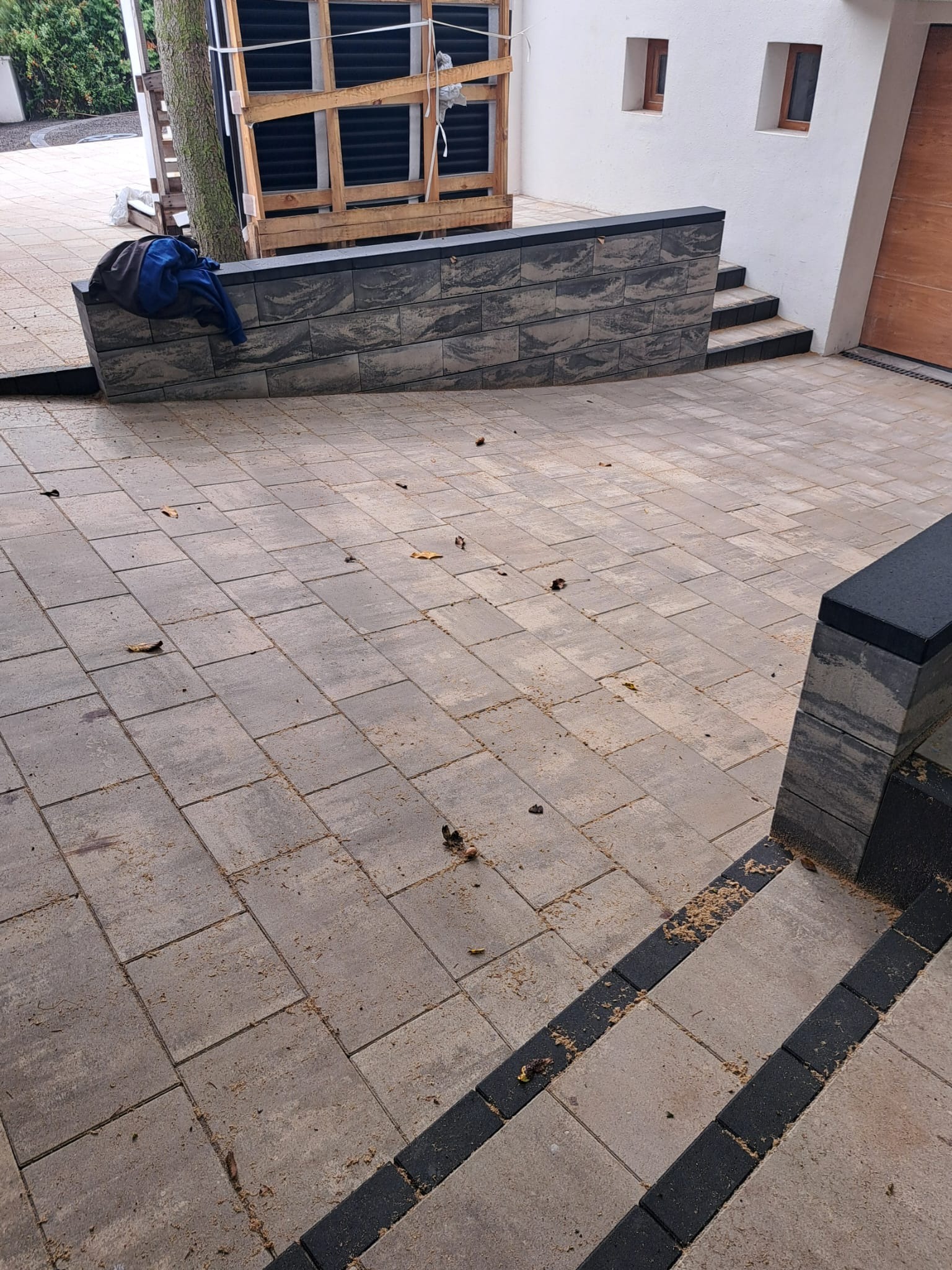 Ułożona kostka brukowa w odcieniach szarości tworzy wzór na podjeździe. Widoczne schody z kostki i fragment ściany budynku z oknami. Na murku leży niebieska kurtka.