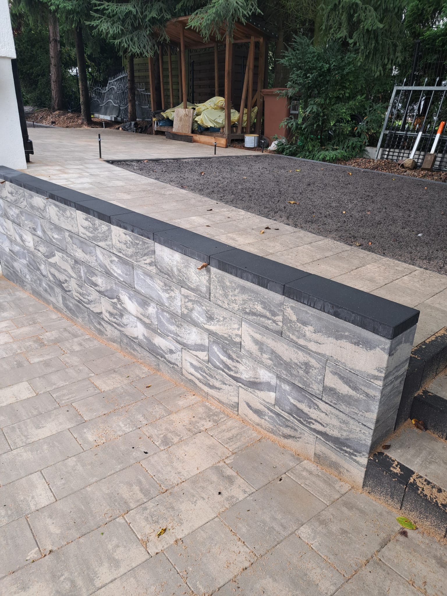 Mur z kostki dekoracyjnej z czarnym wykończeniem, otoczony brukowaną ścieżką i przygotowanym terenem pod nasadzenia. Widoczne narzędzia ogrodnicze w tle.