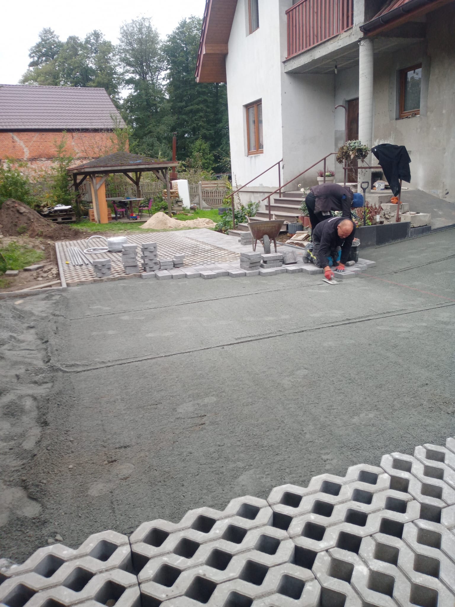 Układanie ażurowej kostki brukowej na podjeździe przed domem. Dwóch brukarzy w trakcie pracy, widoczne narzędzia i przygotowane materiały. Częściowo ułożona nawierzchnia.