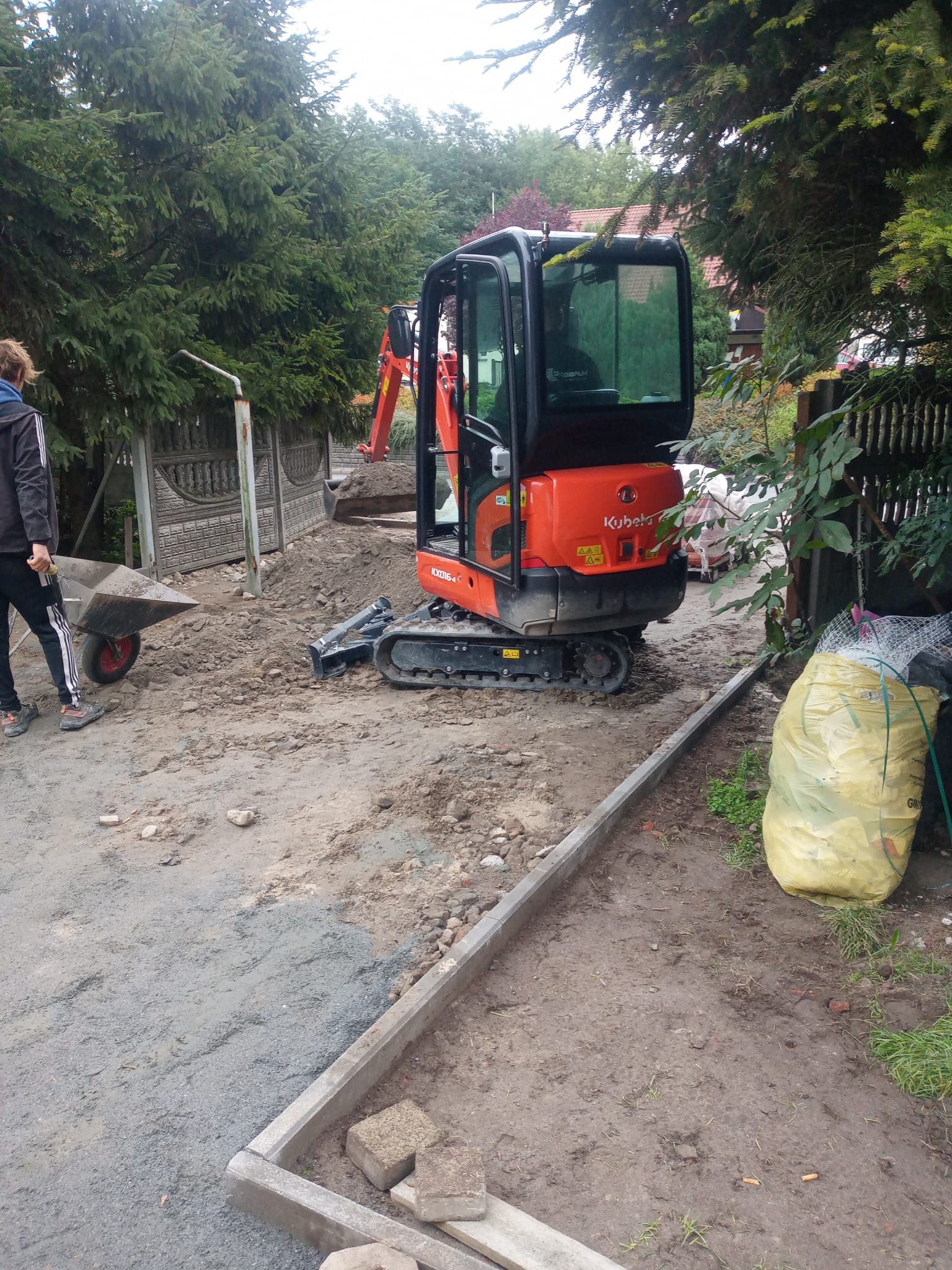 Pomarańczowa mini koparka Kubota na placu budowy, obok wózek i worki z odpadami, w tle ogrodzenie i zieleń. Przygotowanie terenu pod kostkę.