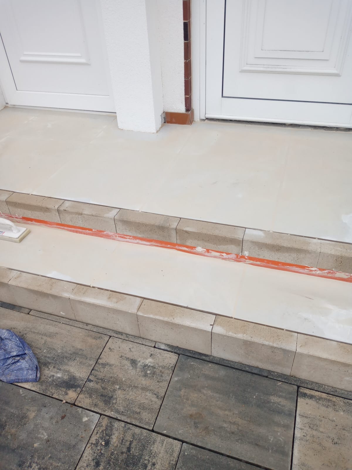 Wykończone płytki na schodach wejściowych z obrzeżami z kostki, widoczne narzędzia i fragment kostki brukowej. Jasne płytki kontrastują z ciemniejszą kostką.