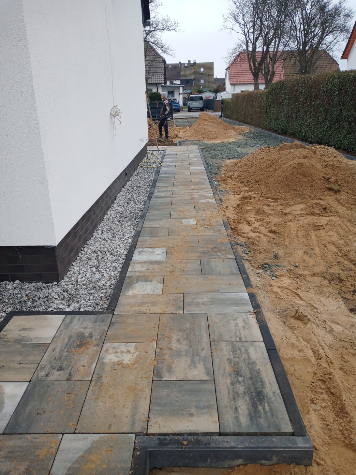 Ścieżka z kostki brukowej w trakcie układania, widoczne przygotowane podłoże z piasku i żwiru, pracownik w tle. Kostka w odcieniach szarości i beżu.