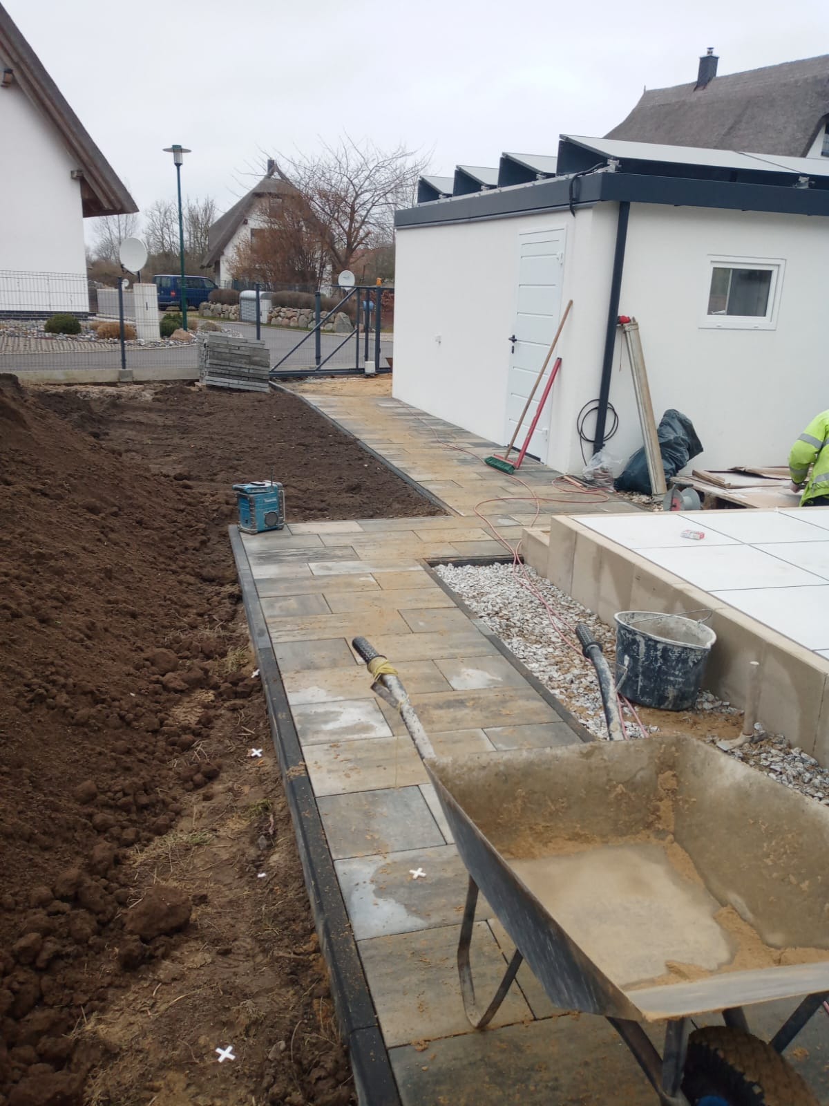 Układanie kostki brukowej: widok na ścieżkę z kostki, obrzeża i przygotowany teren pod trawnik. W tle budynek z dachem krytym strzechą i biały budynek gospodarczy.