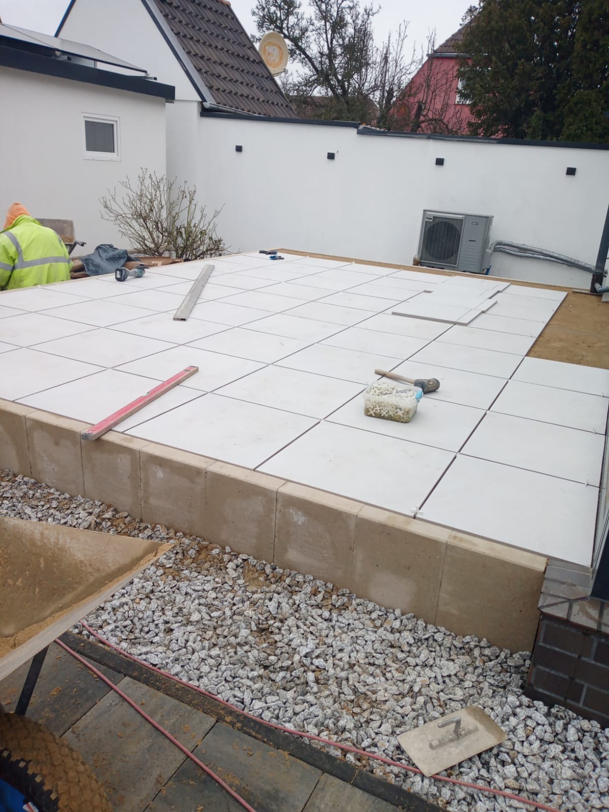 Białe płytki tarasowe układane na podwyższonym fundamencie, widoczne narzędzia i częściowo ułożona powierzchnia. W tle budynek i ściana.
