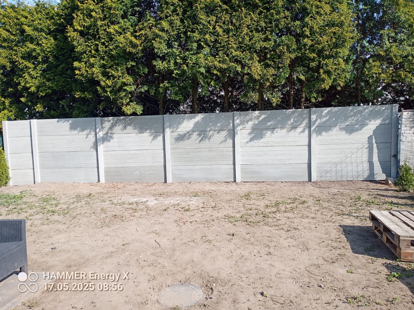 Nowoczesny, prefabrykowany płot betonowy o fakturze drewna, oddzielający posesję od zieleni drzew. Widoczne elementy montażowe i surowy teren przed ogrodzeniem.