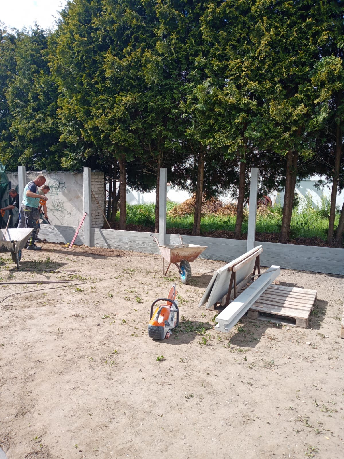 Montaż ogrodzenia panelowego z betonową podmurówką. Widoczne słupki, panele, taczki i piła do betonu. W tle rząd drzew.